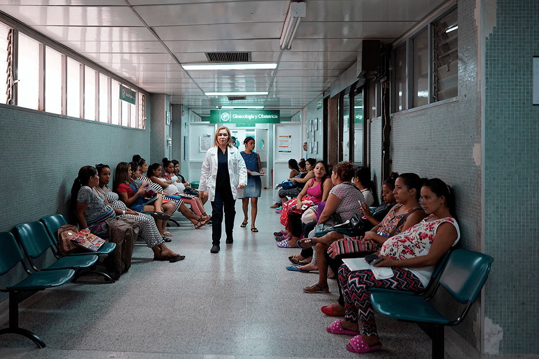 Health care worker walking down an overcrowded hallway lined with women waiting to be seen in a hospital in Colombia. 