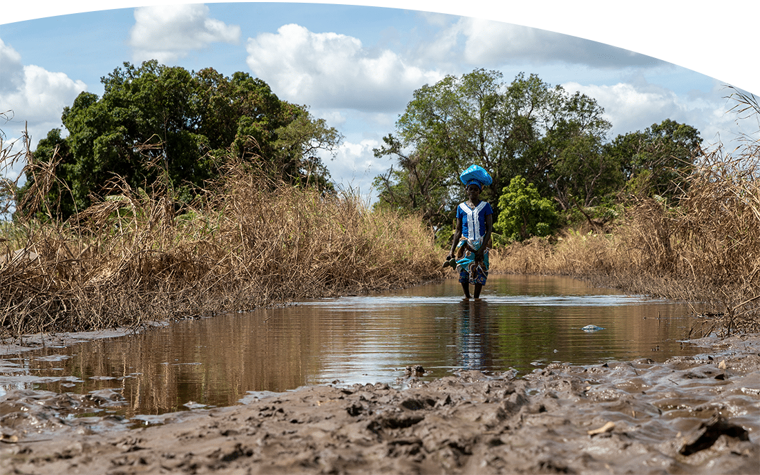 Woman in Mozambique walking on a flooded path carrying items on her head and her shoes in her hands.