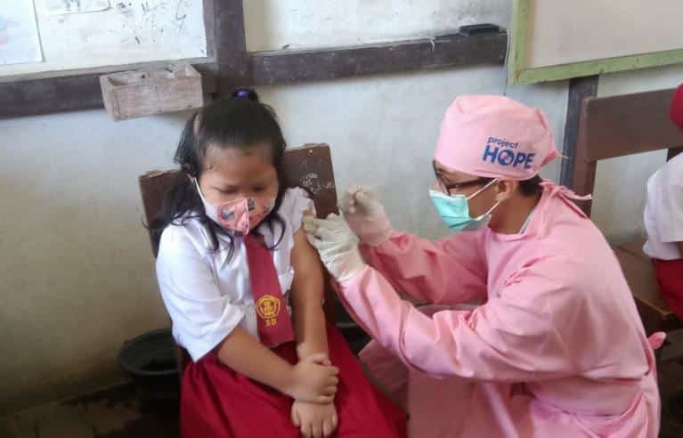 School girl in a mask receives a vaccine by a doctor in pink scrubs.