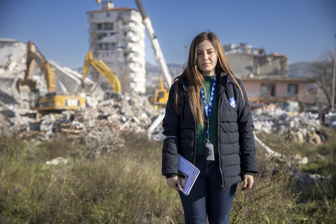 woman standing in front of destroyed buildings in Turkiye, looking into camera with sad expression.