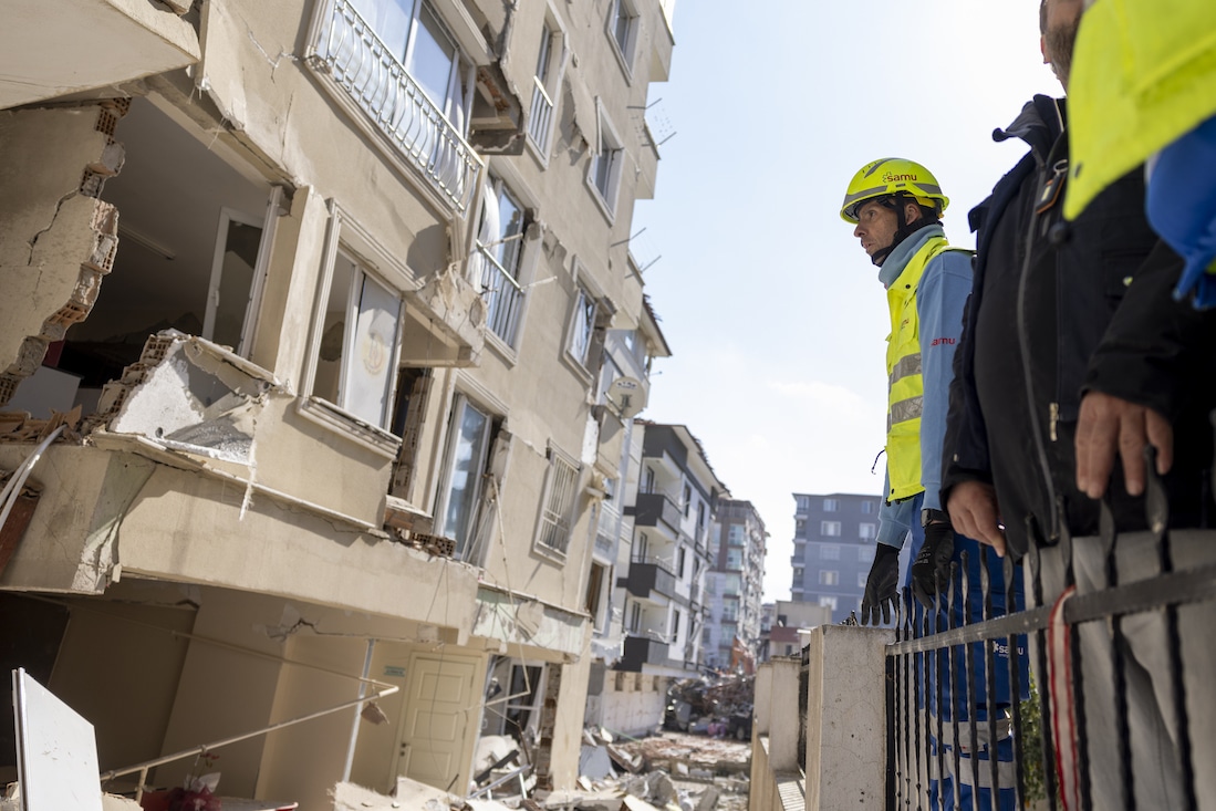 men in helmets look on at destroyed building in Turkiye.