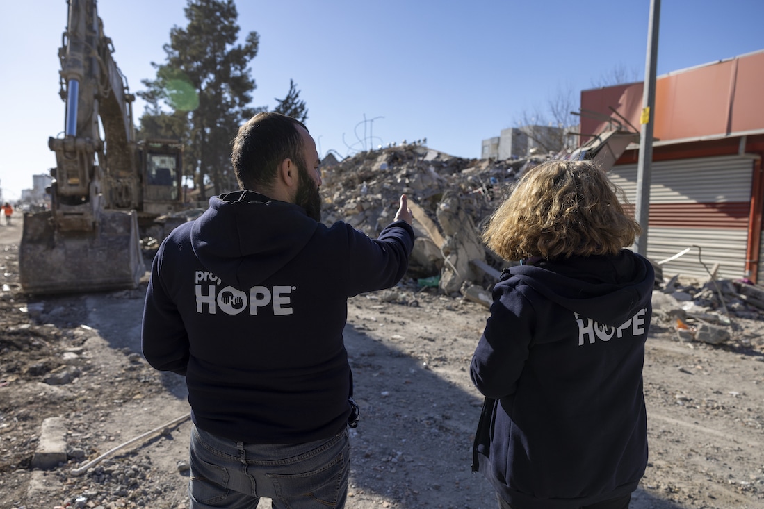 Visible behind the backs of two people wearing Project HOPE sweaters as they discuss rubble in front of them.