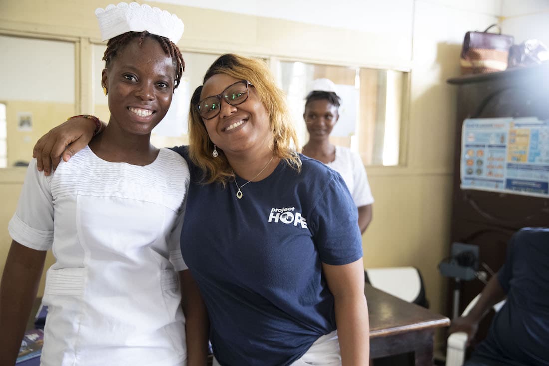a woman in a navy shirt stands next to a nurse in a white dress in a hospital. They are both smiling