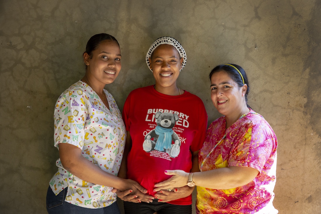Two medical staff stand next to a pregnant woman, holding her stomach. They all smile at the camera