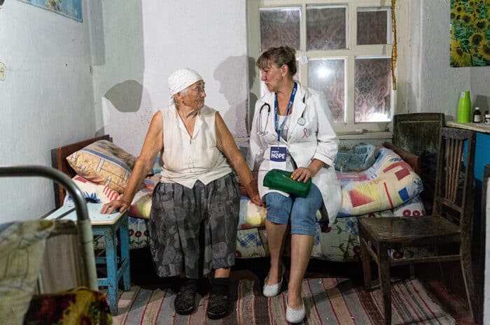 A nurse sits with an elderly patient on a bed