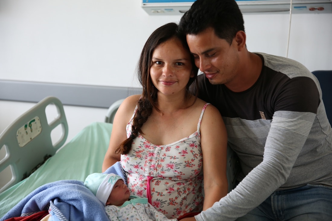 Latin mother and father sitting in hospital bed holding newborn baby. Mother is looking at the camera.