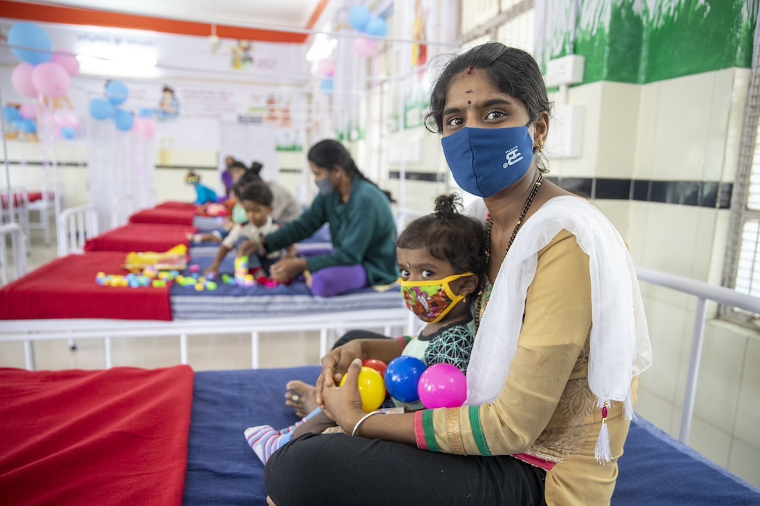 South Asian mother in a mask holding young child, also in a mask, while they sit on a medical bed.