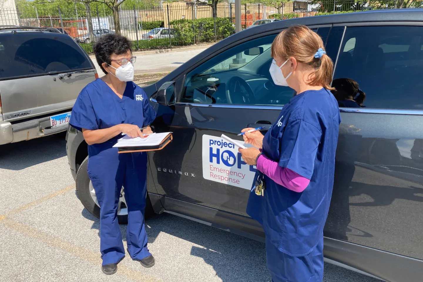 Two medical staff in scrubs and facemasks outside of cars discussing COVID-19