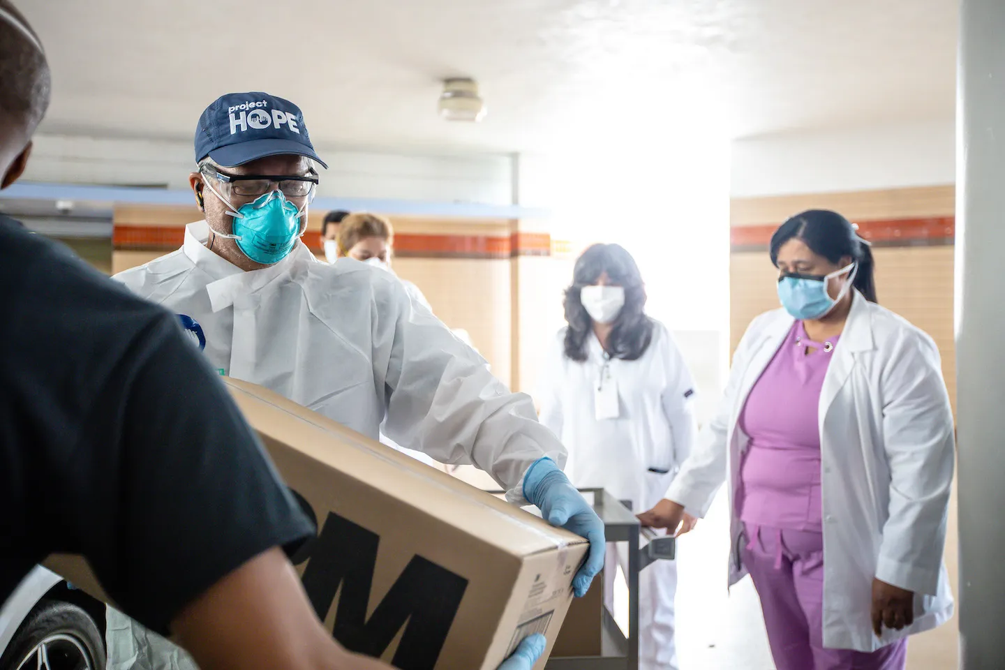 Man in full PPE gear carrying a box of medical supplies