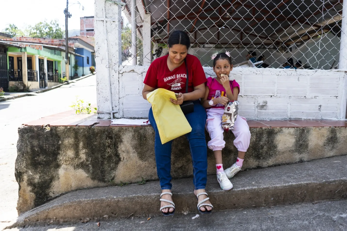 A mother and daughter sitting together on the side of a street eating snacks.