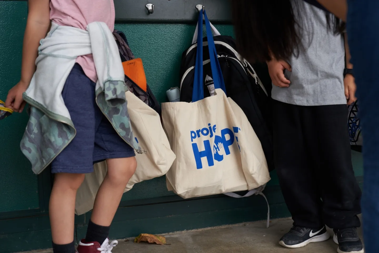 image of waist and below of two young boys standing in a hallway with Project HOPE totes handing on hooks next to them.
