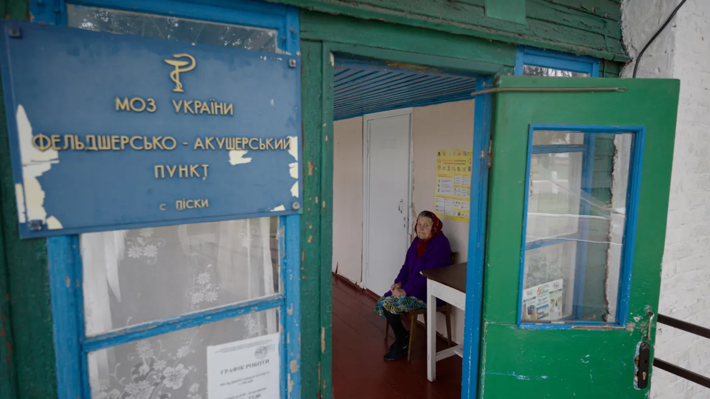 older woman sits inside a building. We see her from outside the front door