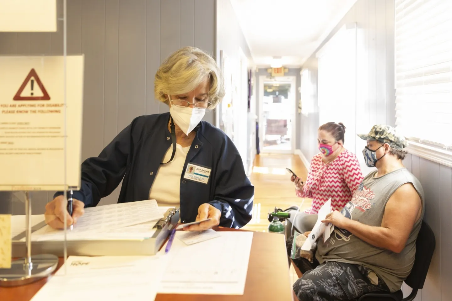A medical staffer documenting information at a clinic while two people wearing facemasks wait in the lobby.