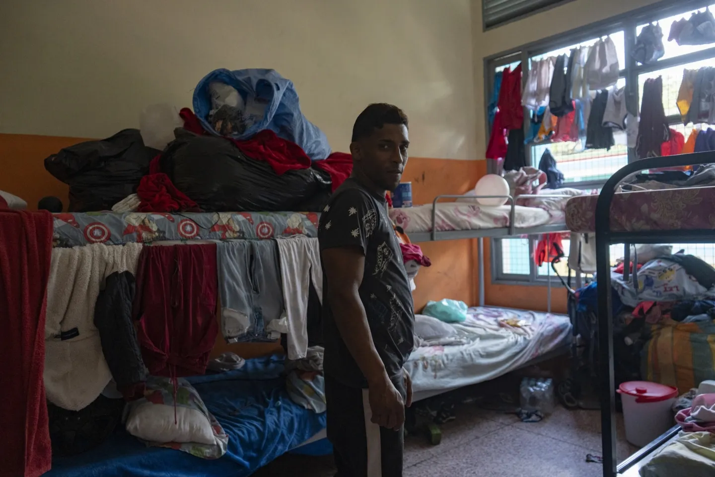man stands in the middle of a room of three bunkbeds with clothes hanging from the rails and windowpanes where he and his family is sheltering after the earthquake in Venezuela