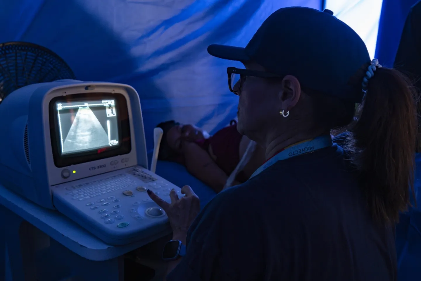 blue-lit photo of woman inside medical tent observing an ultrasound of pregnant woman laying next to her.