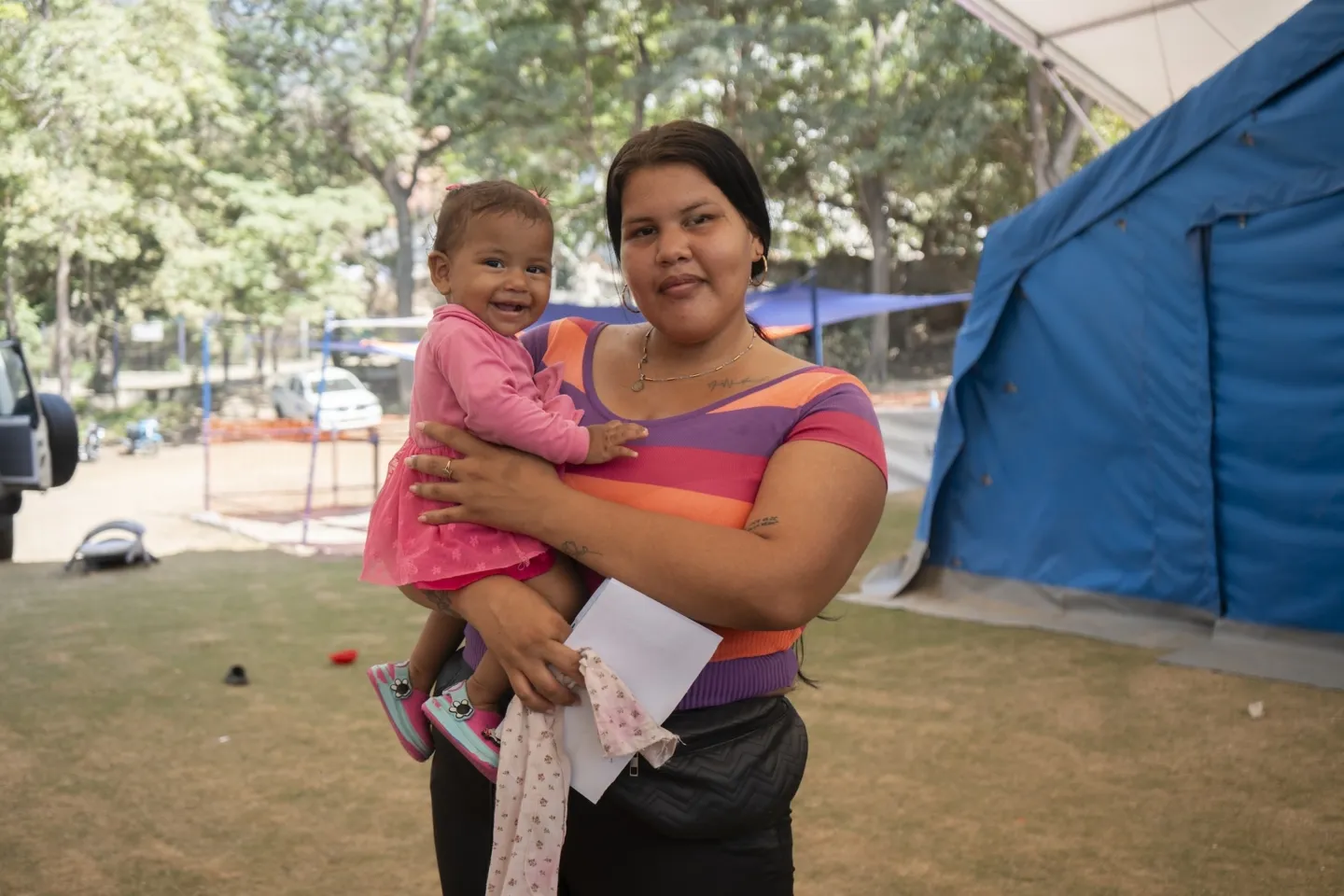 woman in colorful striped shirt holds young daughter in a pink dress outside a pop-up medical tent in Venezuela