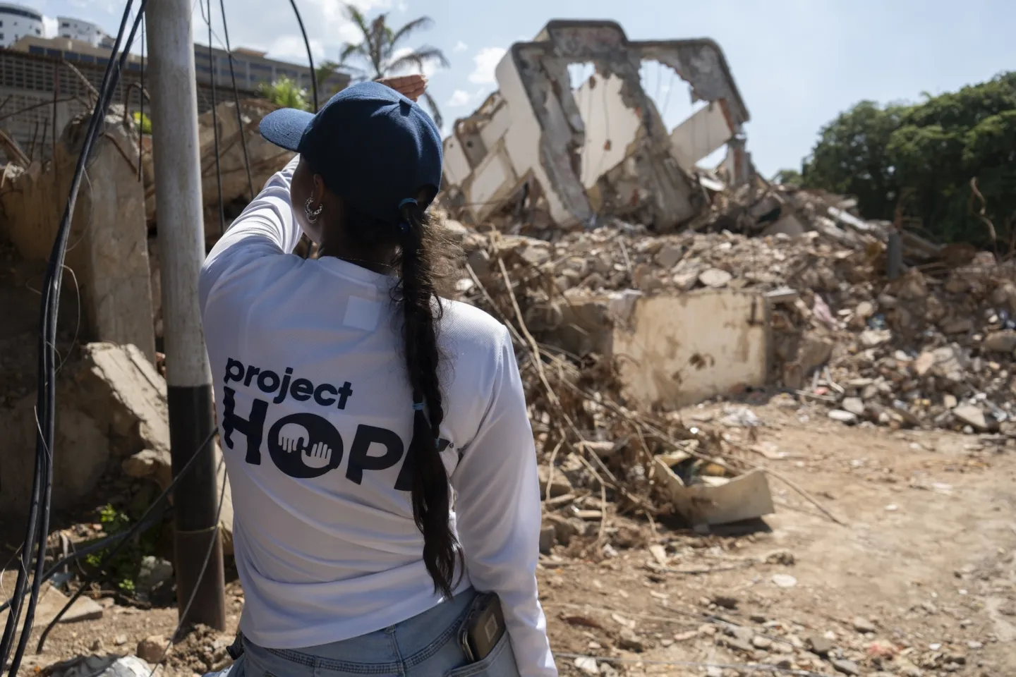 woman in baseball hat and Project HOPE shirt faces destroyed building in Venezuela
